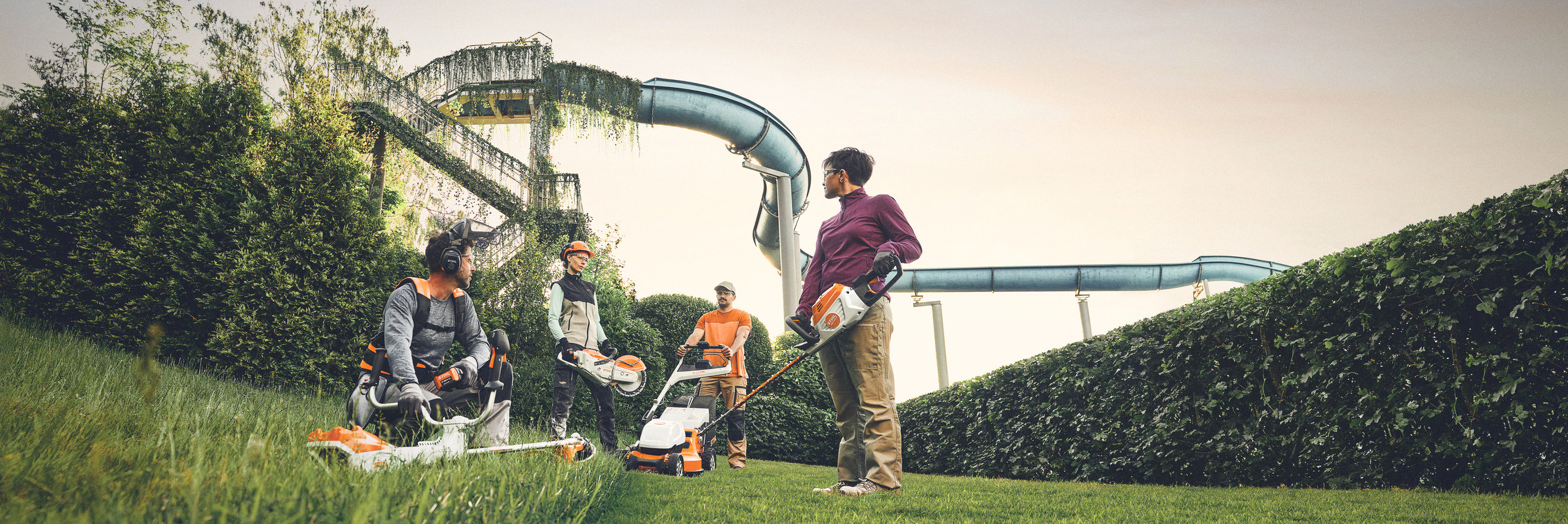 En grupp människor arbetar med STIHL trädgårdsredskap på ett grönt område med en stor häck och vattenrutschbanor i bakgrunden. De är utrustade med röjsågar, gräsklippare och skyddsutrustning.