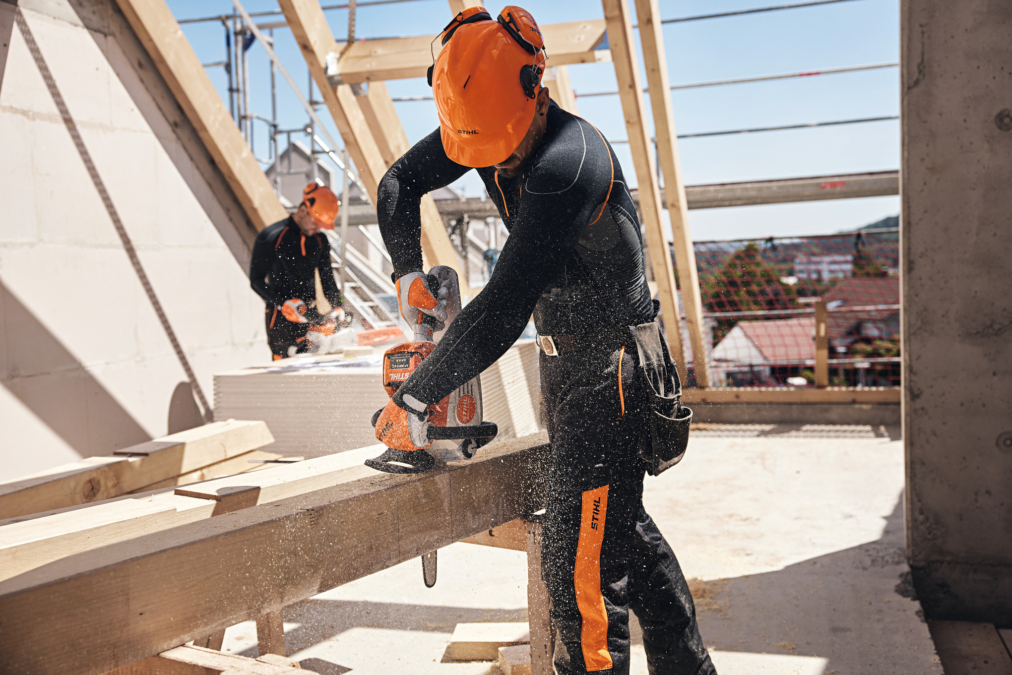 Ett STIHL-proffs på ett loft sågar en träbalk med en batteridriven motorsåg, medan en annan arbetare använder en batteridriven motorsåg i bakgrunden