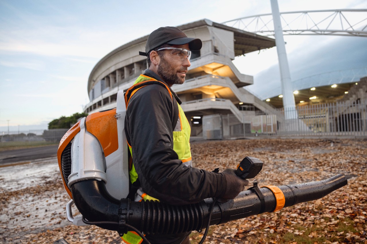 Sent på kvällen röjer en person löv och skräp från en stadion med hjälp av den sladdlösa lövblåsen ALLPRO STIHL BRA 600.