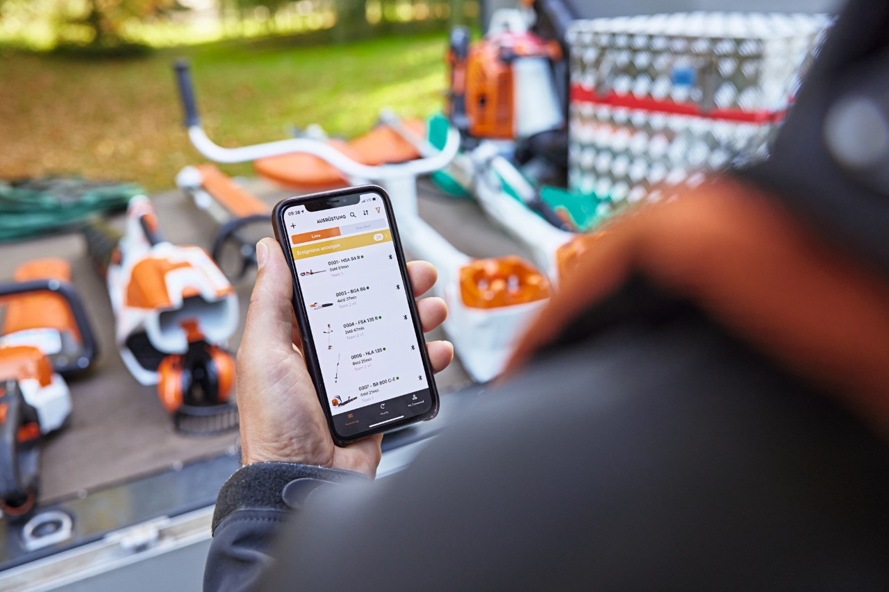 En person som står framför lastytan på ett flakfordon med olika redskap från STIHL. Personen håller en smartphone med STIHL connected-appen öppen. 