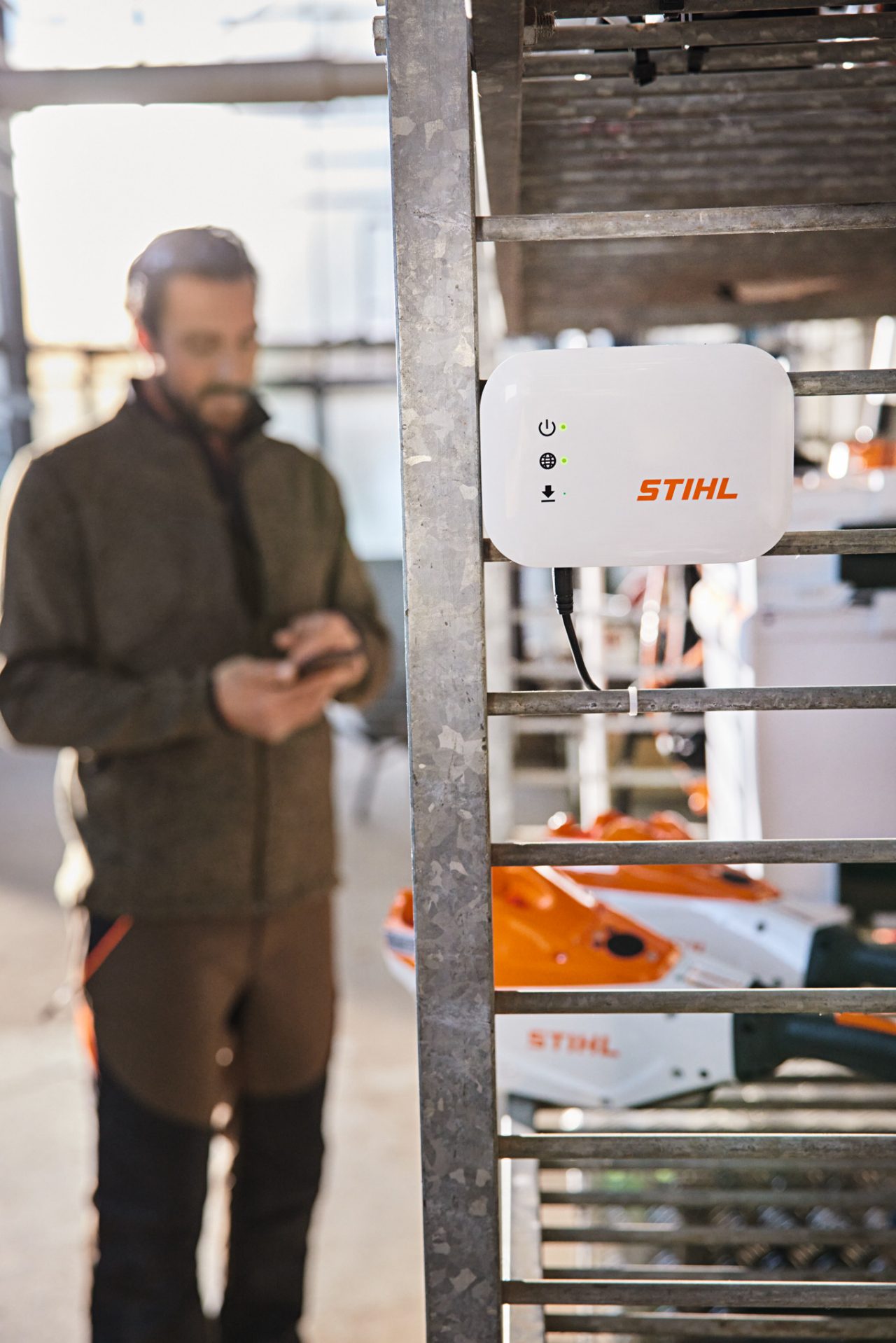 En STIHL connected Box hänger på en verkstadshylla. Med en trädgårdsmästare och landskapsarkitekt i bakgrunden som använder en smartphone. 