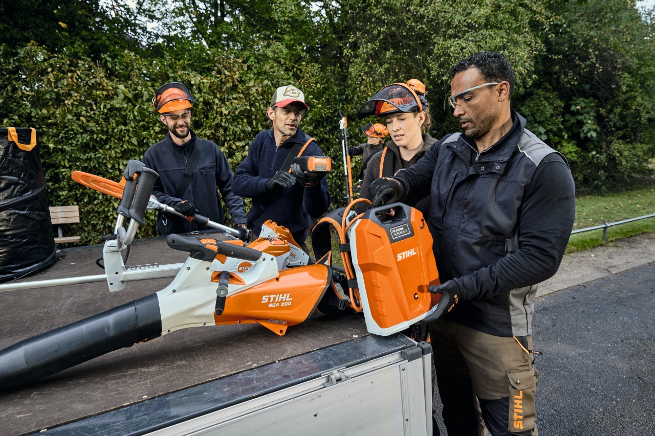 Tre män och en kvinna med STIHL skyddsutrustning står på ett fordonsflak med maskiner från STIHL såsom STIHL BGA 200 batteridriven lövblås och STIHL AR 2000 L ryggburet batteri 