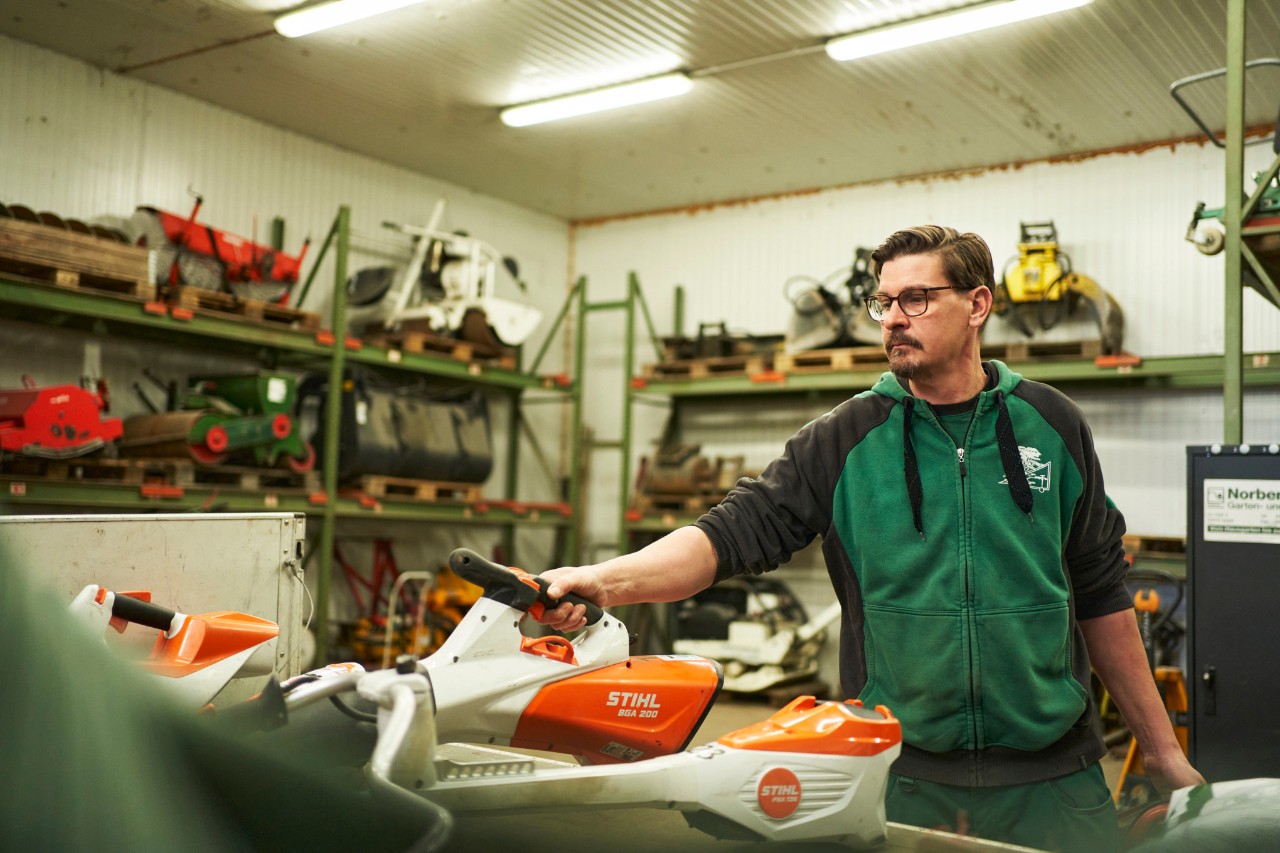 En man i en verkstad står bakom en arbetsyta med STIHL batteridrivna maskiner och sträcker sig efter en STIHL batteridriven lövblås BGA 200
