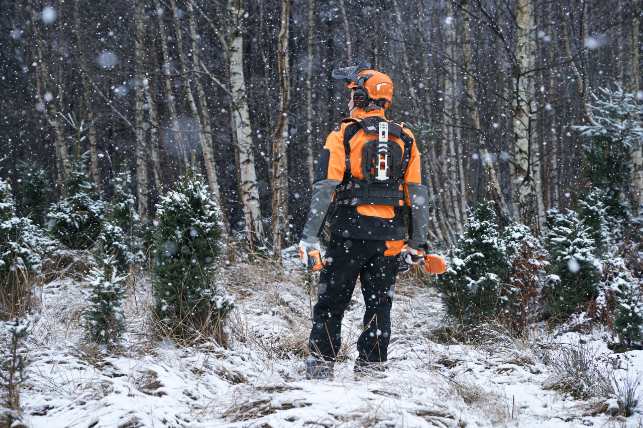 En skogsarbetare med STIHL skyddsutrustning med ett modulärt selsystem och batteridrivna maskiner för underhåll av unga trädbestånd i en snöig skog, sedd bakifrån