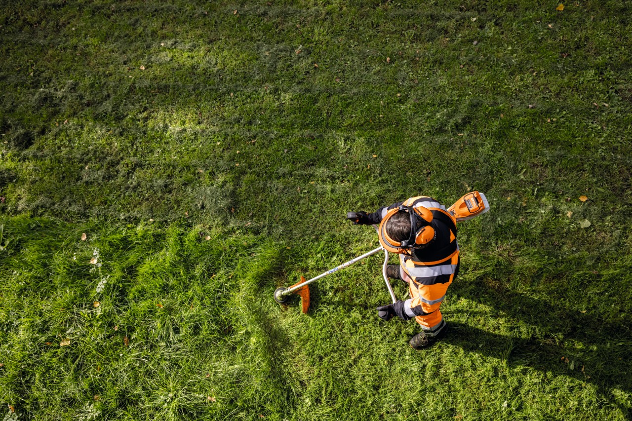 Trädgårdsmästare och landskapsarkitekt i orange arbetskläder klipper högt gräs med STIHL FSA 135 batteridriven röjsåg