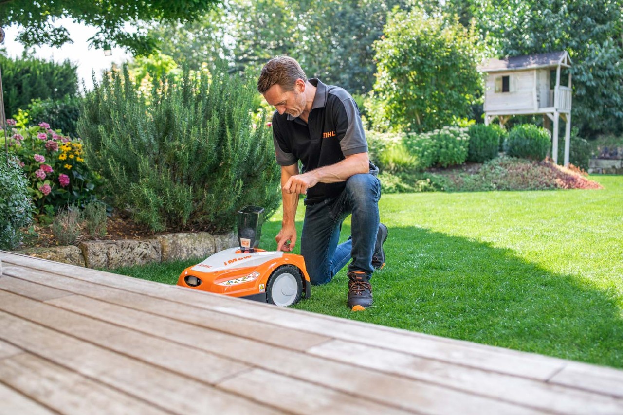 En man knäböjer på gräsmattan bredvid en STIHL iMOW® robotgräsklippare i en trädgård med buskar och altandäck