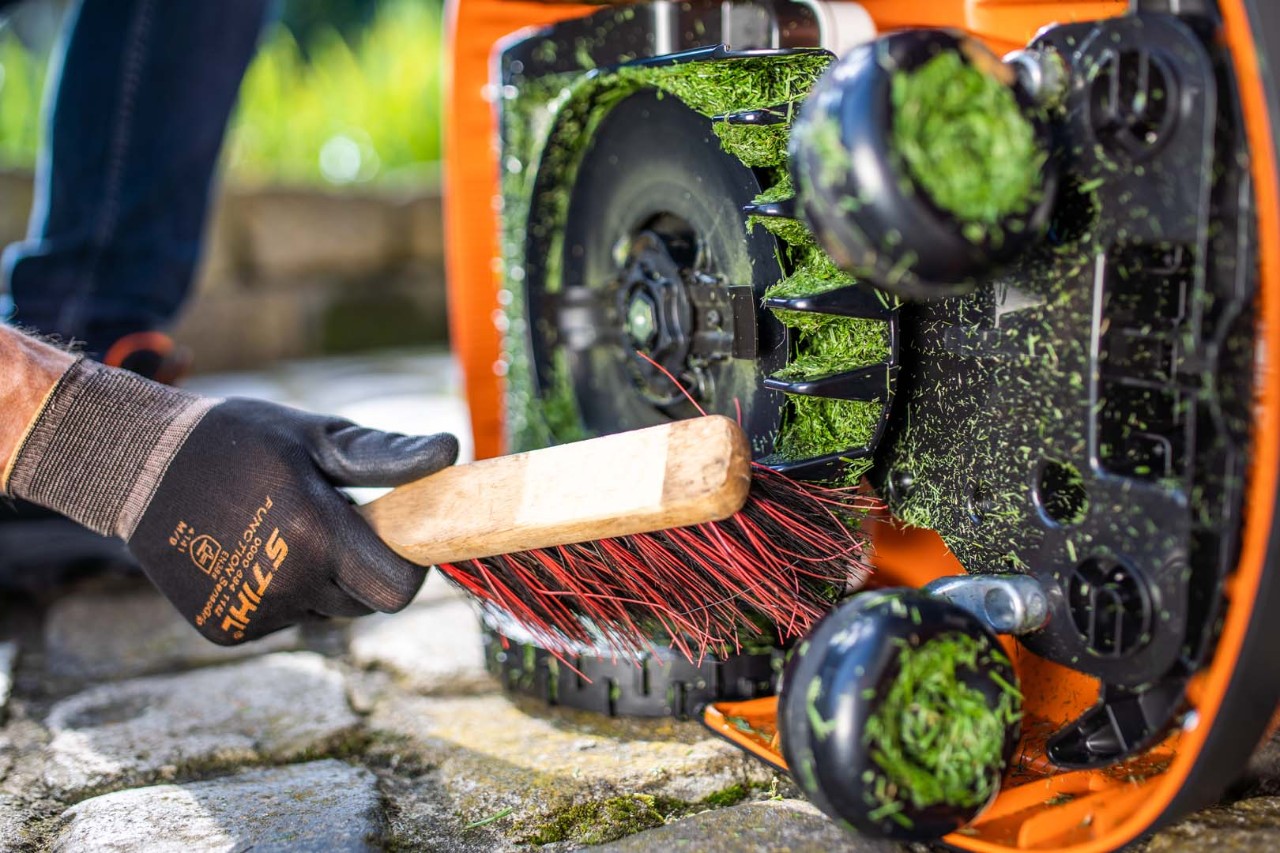 Gräsklippaggregatet på en STIHL iMOW® robotgräsklippare vänd på sidan som rengörs med en borste av en person med handskar