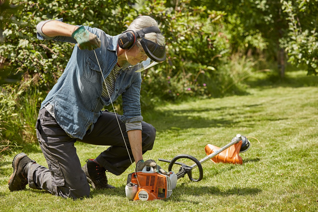 En kvinna som bär hörsel- och ansiktsskydd är på väg att starta en STIHL bensindriven trimmer FS 56 R på en grön tomt