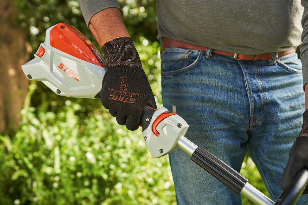 En man som bär skyddshandskar trycker på gasspaken på STIHL FSA 60 batteridriven trimmer för att starta den