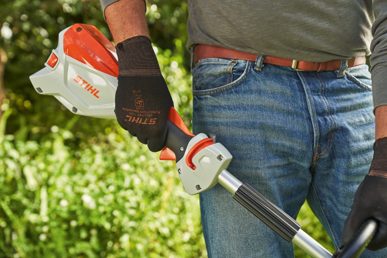 En man som bär skyddshandskar släpper gasspaken på STIHL FSA 60 batteridriven trimmer för att stänga av den