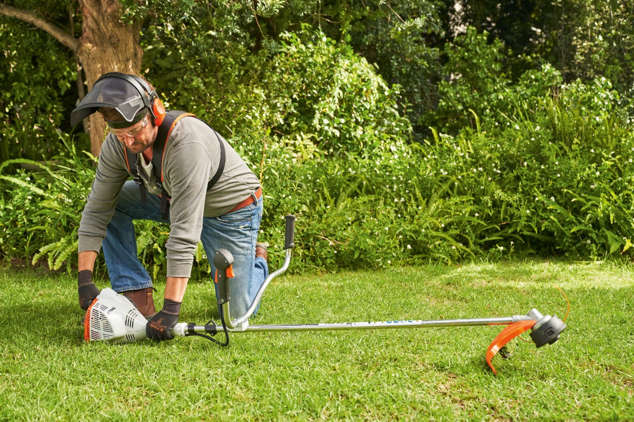 En man som bär hörsel- och ansiktsskydd är på väg att starta en STIHL FS 56 R bensindriven trimmer i en trädgård