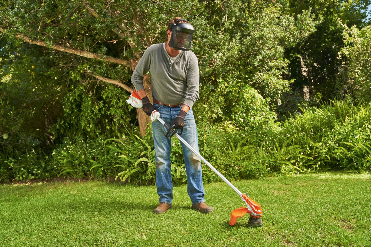 En man med ansiktsmask klipper gräsmattan i en trädgård med STIHL FSA 60 batteridriven trimmer