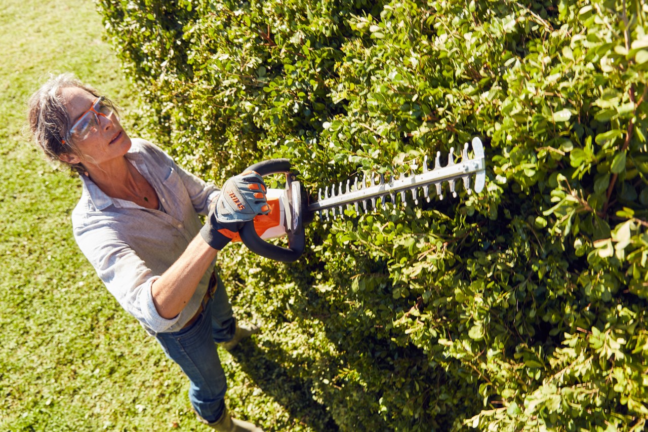 Kvinna med skyddsglasögon klipper en hög häck med en STIHL HSA 56 batteridriven häcksax