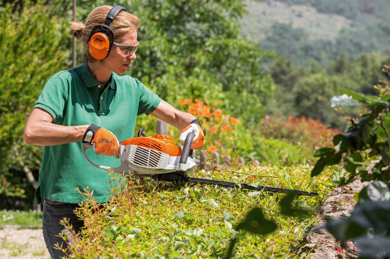 En kvinna som bär hörselskydd och skyddsglasögon klipper en häck med en STIHL bensindriven stånghäcksax HS 46