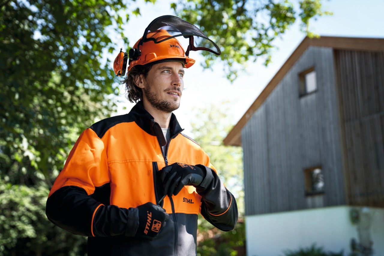 En man med orange och svart skyddsutrustning från STIHL, inklusive hjälm och handskar, tittar självsäkert på oss från bilden med ett hus och träd i bakgrunden 