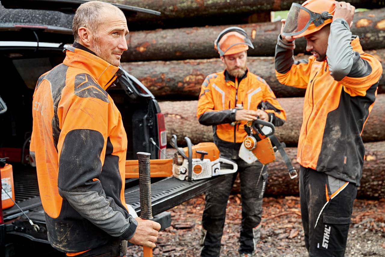 Tre män i STIHL skyddskläder gör sig redo att använda en STIHL motorsåg framför en vedhög