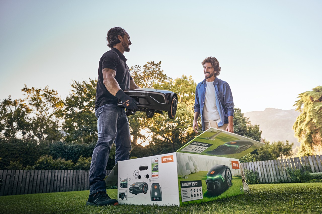 En STIHL-återförsäljare står i en kunds trädgård med en iMOW® EVO i handen och iMOW®-lådan framför sig, redo att hjälpa till med installationen